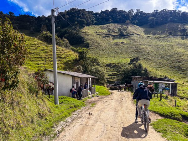 Bicyclists ride along a dirt path, flanked by green hills and rustic houses in Carbonera Valley, Quindío, Colombia.