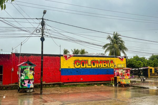 Colorful Colombia mural on a red wall with yellow and blue stripes, surrounded by palm trees and street vendors in Leticia.