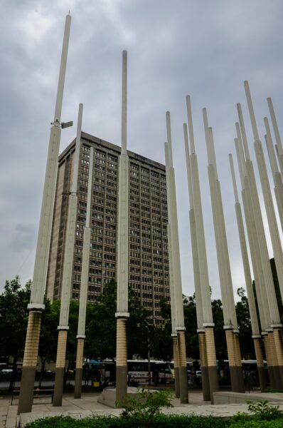 Tall, white columns rise toward a cloudy sky in Parque de las Luces, with a modern building visible in the background.