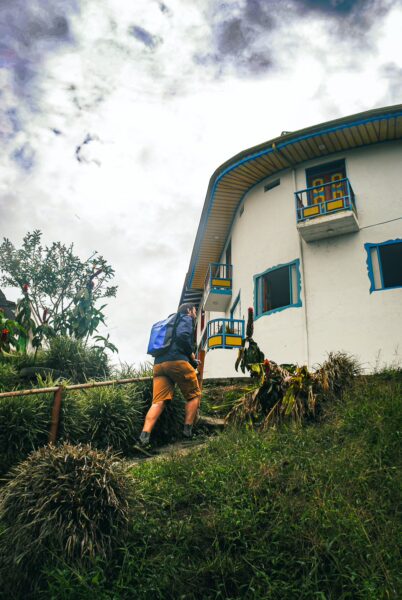 A traveler ascends a grassy path to a white building with blue accents in Salento, Quindío, under a cloudy sky.