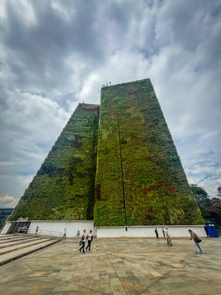 Two tall, tapered green facade towers rise over a paved plaza in central Medellín under a cloudy sky, with a few people walking below