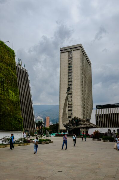 Visitors gather around the Monumento a la Raza in Medellín, with modern buildings and a cloudy sky in the background.