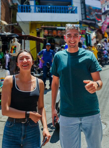 Two smiling people stand in a vibrant street of Comuna 13, surrounded by bustling vendors and colorful murals.
