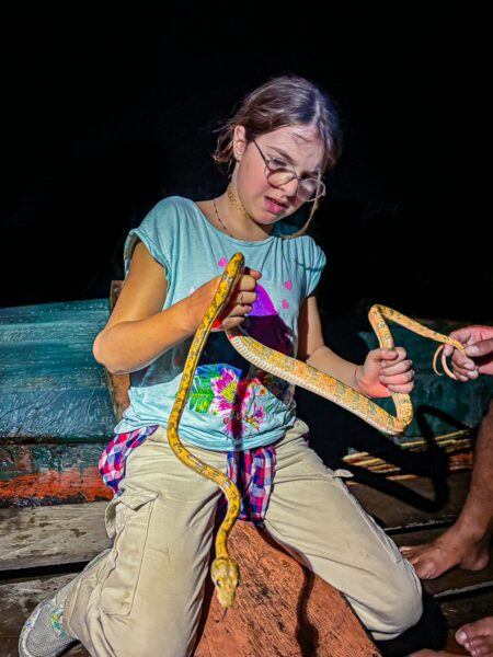 A young girl in a colorful shirt carefully holds a yellow snake inside Gamboa Nature Reserve, Loreto, Peru.