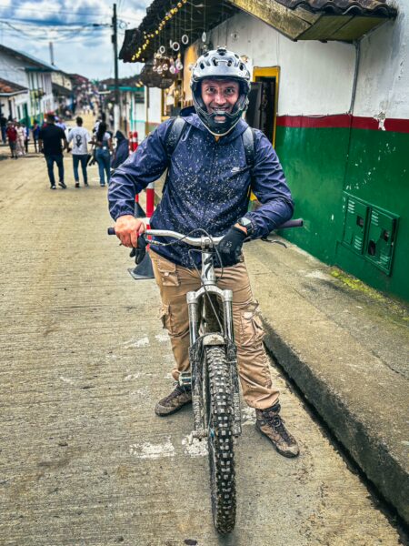 Biker equipped with a helmet and camera smiles while posing with a mountain bike on a colorful street in Salento, Quindío, Colombia.