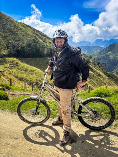 Biker in protective gear stands beside a mountain bike, with lush green hills of Carbonera Valley, Quindío, Colombia, in the background.