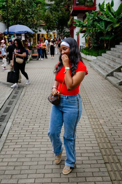 A young woman in a red top and jeans smiles while walking through the vibrant streets of Pueblito Paisa in Medellín.