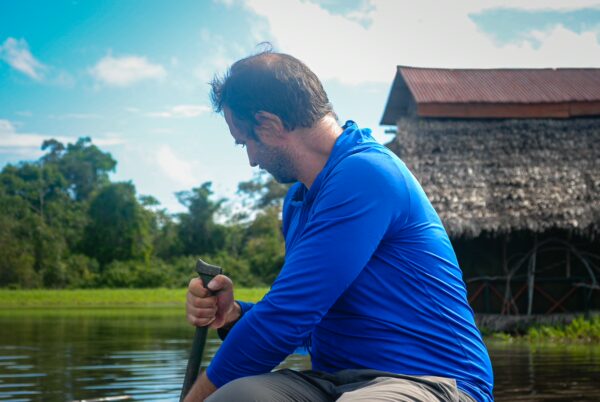 Paddler in a blue shirt navigates serene waters at Pirana Lake, surrounded by lush greenery and a traditional thatched-roof hut in Gamboa Nature Reserve, Loreto, Peru.