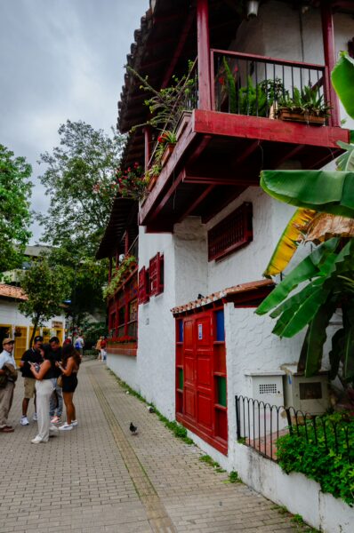 Pueblito Paisa features bright red balconies and white walls along a cobblestone path, surrounded by lush greenery and visitors.