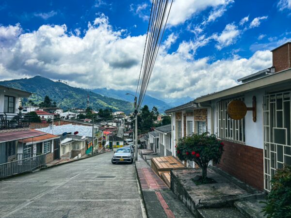 Colorful houses line a quiet street in Salento, Quindio, Colombia, with rolling hills and cloudy blue skies in the background.
