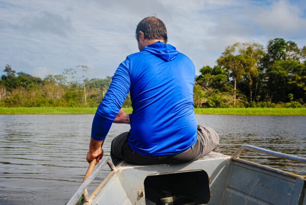 A man in a blue hoodie paddles a boat on Pirana Lake, surrounded by lush greenery in Gamboa Nature Reserve, Loreto, Peru.
