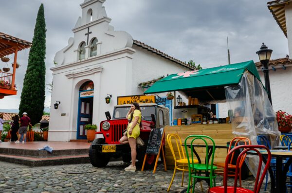 Pueblito Paisa courtyard with a small white chapel, colorful café chairs, and a red jeep under a cloudy sky