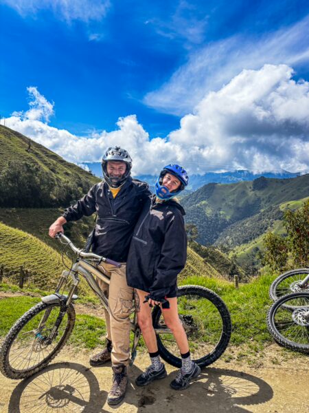 Cyclists pose on mountain bikes in lush Carbonera Valley, Quindio, Colombia, with vibrant green hills and a blue sky backdrop.