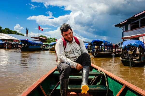 A traveler in a boat adjusts his boots at a bustling dock in Santa Rosa, Amazonas, Peru, with vibrant boats and cloudy skies nearby.