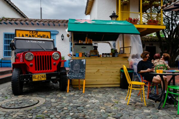 Cobbled plaza at Pueblito Paisa with a café kiosk, colorful chairs, and a vintage red Jeep beside white colonial-style buildings