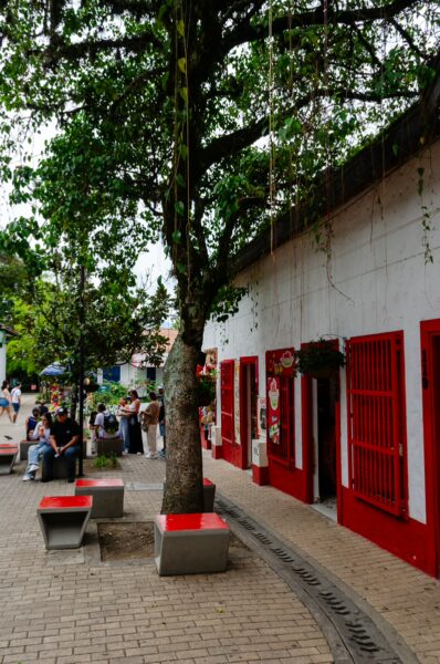 Pueblito Paisa features vibrant red and white buildings, shaded by a large tree, with visitors enjoying the charming atmosphere.