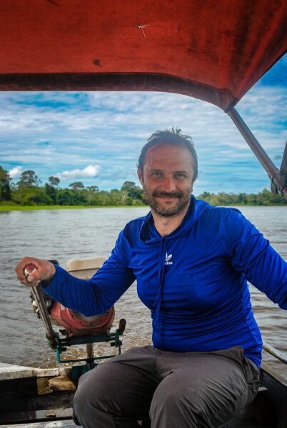 A person in a blue shirt smiles while sitting on a boat, with lush greenery and Pirana Lake visible in the background, Gamboa, Loreto, Peru.
