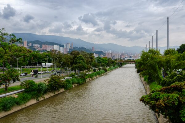 A tranquil river flows through Medellín, flanked by lush greenery and modern buildings under a cloudy sky.