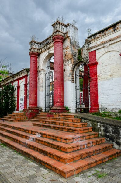 Weathered entrance at San Lorenzo Cemetery in Medellín, with red brick steps, whitewashed walls, and iron gates beneath a cloudy sky