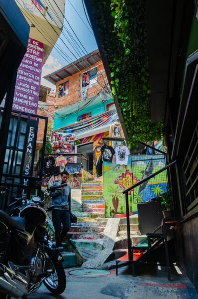 Vibrant street art adorns the colorful stairway in Comuna 13, Medellin, with a backdrop of bustling market stalls and greenery.