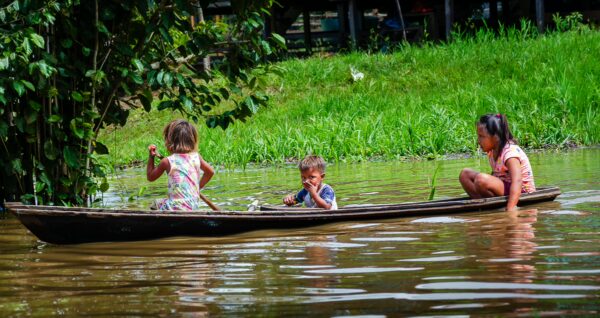 Children play in a wooden canoe among lush greenery in Gamboa Nature Reserve, Loreto, Peru. Reflections shimmer in the water.