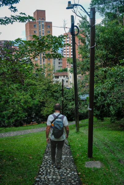 A man walks along a stone pathway surrounded by greenery and urban buildings in Pueblito Paisa, Medellín.