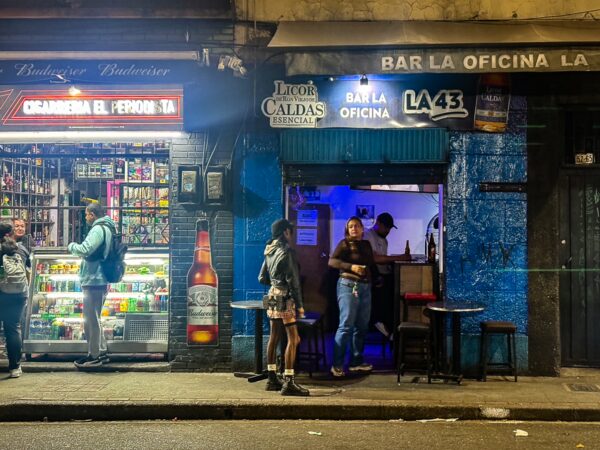 Nightlife street scene on Carrera 42 in Medellín with bar fronts, neon signs, and people standing outside on the sidewalk