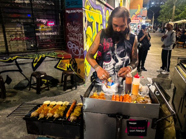 Street food vendor grilling skewers on Carrera 42A in Medellín at night, with graffiti-covered walls and nightlife crowds nearby