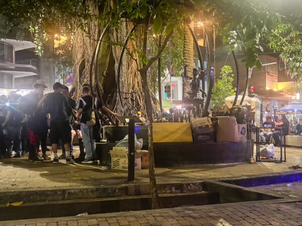 Night scene in a crowded plaza with a large tree, people gathered around, and street vendors under warm lights