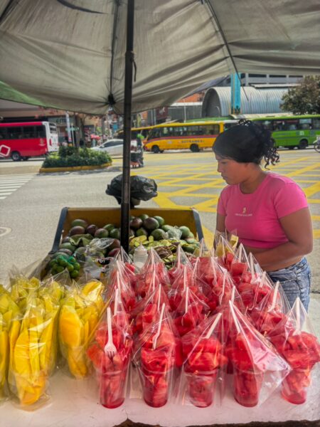 Local vendor selling colorful fruits, including yellow mango and red watermelons, at a bustling street market in Medellín, Colombia.