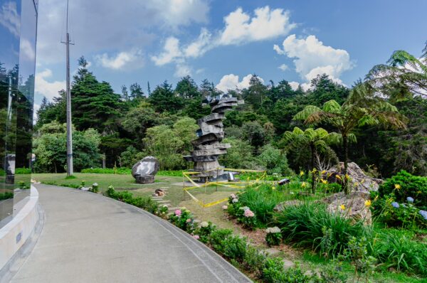 Arvi Park features a stone sculpture surrounded by lush greenery and colorful flowers under a blue sky in Medellín, Colombia.
