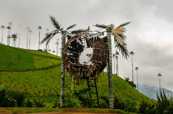 Cocora Valley's iconic wax palm stands beside a rustic sculpture, framed by lush green hills and a cloudy sky in Quindío, Colombia.