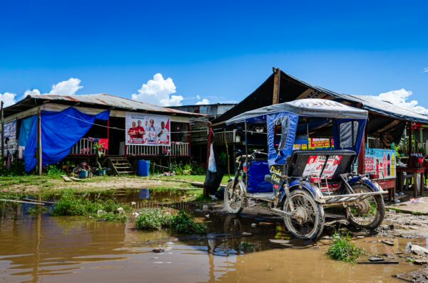 Colorful storefronts and a bicycle taxi are reflected in the muddy water by the vibrant landscape of Santa Rosa, Amazonas, Peru.
