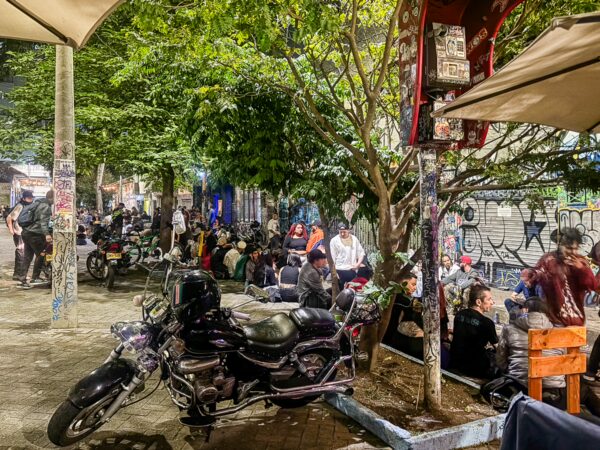 Vibrant streets of Medellín at night, filled with people socializing under lush trees and colorful graffiti. Motorcycles parked nearby.