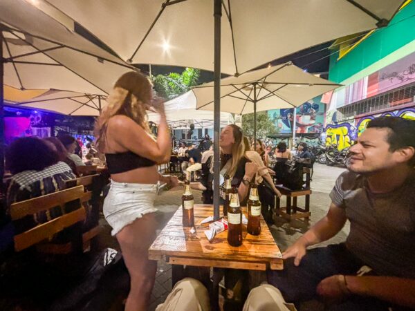 Nighttime street bar in Medellín with people seated under large umbrellas, bottles on a wooden table, and graffiti-covered walls nearby