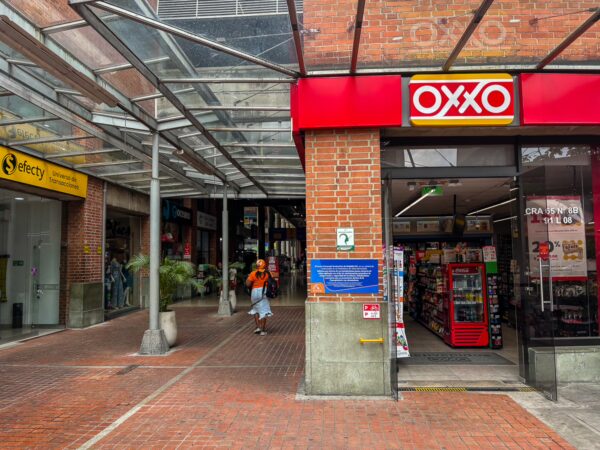 A brick building entrance features an OXXO convenience store under a transparent rooftop, with a pedestrian walking by in Medellín.