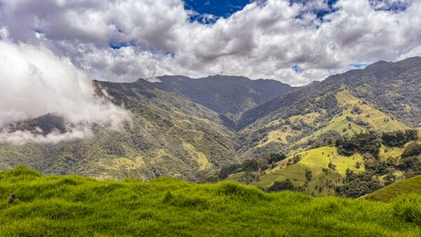 Lush green hills and towering mountains under a bright blue sky with fluffy clouds in Carbonera Valley, Quindío, Colombia.