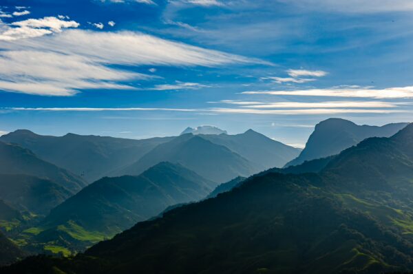 Majestic mountains rise in layers under a blue sky, with soft clouds drifting over the Carbonera Valley in Quindío, Colombia.