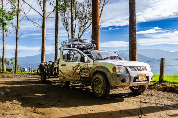 A dusty vehicle parked under tall trees, with rolling green hills and mountains visible in the background of Carbonera Valley, Quindio, Colombia.