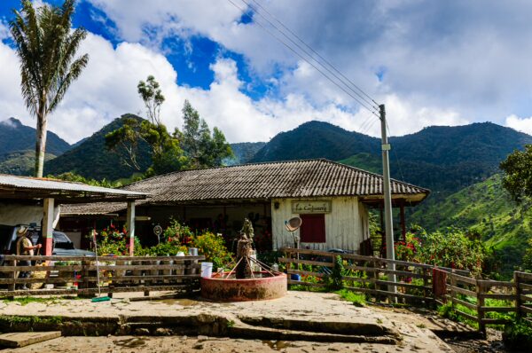 Quaint house surrounded by lush green mountains and vibrant plants in Carbonera Valley, Quindío, Colombia. Sunny skies enhance the scene.
