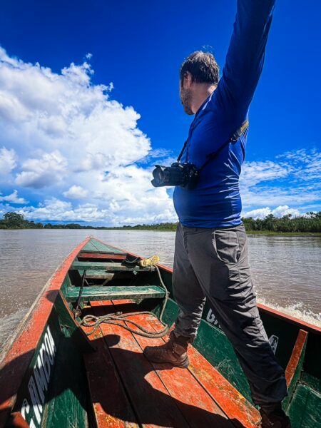A man stands on a wooden boat with a camera, surrounded by lush greenery and a wide river under a blue sky in Santa Rosa, Amazonas, Peru.