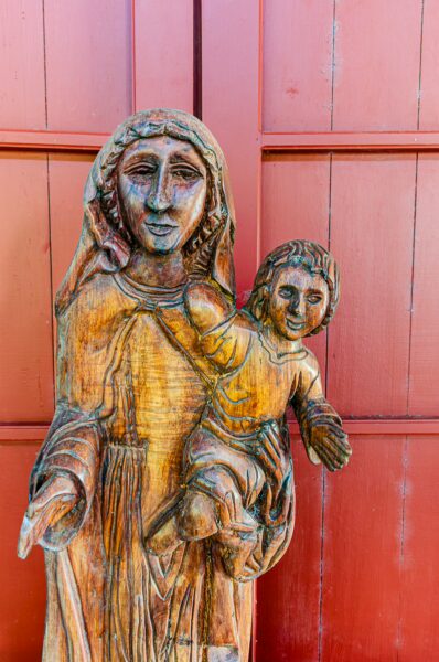 Wooden sculpture of a mother and child, richly detailed with warm tones, set against a vibrant red wall in Puerto Nariño, Amazonas, Colombia.