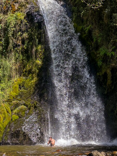 A person stands in the clear water beneath a cascading waterfall surrounded by lush greenery in Carbonera Valley, Quindío, Colombia.