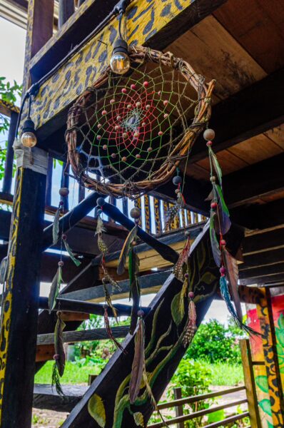 Colorful dreamcatcher hangs above wooden stairs in Puerto Narino, Amazonas, Colombia, with vibrant murals and lush greenery visible.