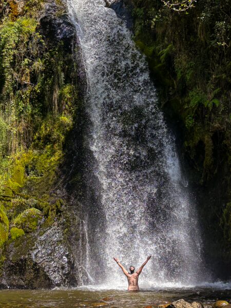 A person stands joyfully under a waterfall, surrounded by lush greenery in Carbonera Valley, Quindio, Colombia.