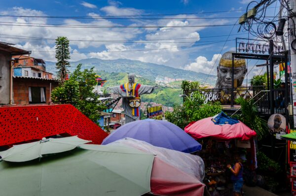 Colorful umbrellas shade a vibrant market in Comuna 13, Medellín, with lush hills and impressive murals in the background.