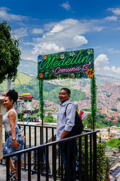Colorful entrance sign to Comuna 13 in Medellín, Colombia, with people enjoying the vibrant atmosphere and scenic hills in the background.