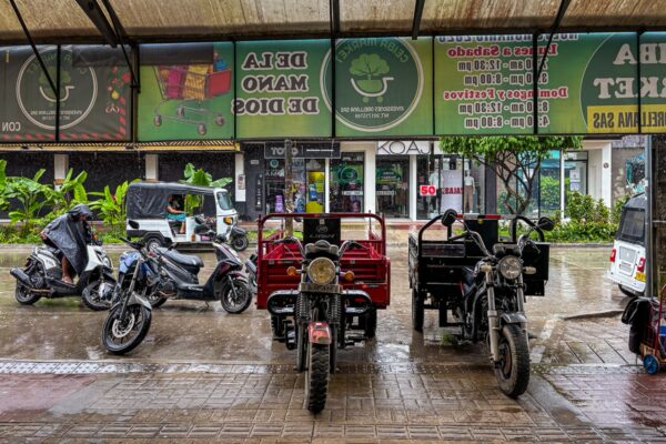 Rain-soaked street scene in Leticia, Amazonas, Colombia, featuring parked motorcycles and three-wheeled vehicles under an awning.