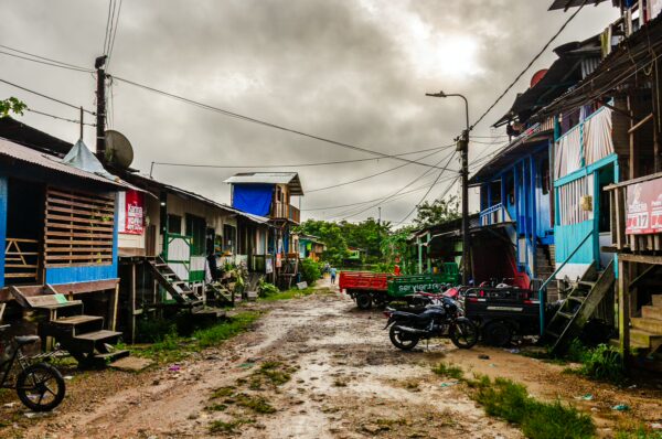 Colorful wooden houses line a muddy street in Leticia, Amazonas, Colombia, under a dramatic, cloudy sky.