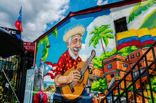 Vibrant mural featuring a smiling musician in a colorful shirt, surrounded by tropical plants and a lively Medellín neighborhood.
